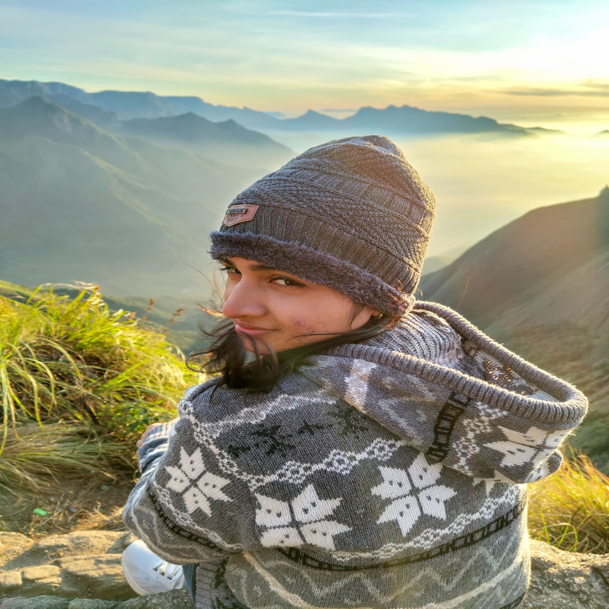 Person in a knitted winter hat and patterned sweater sitting on a mountain ridge during sunrise, overlooking mist-covered valleys.