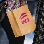 Person carrying a branded shopping bag with a bold logo while walking, alongside a black handbag and patterned clothing.