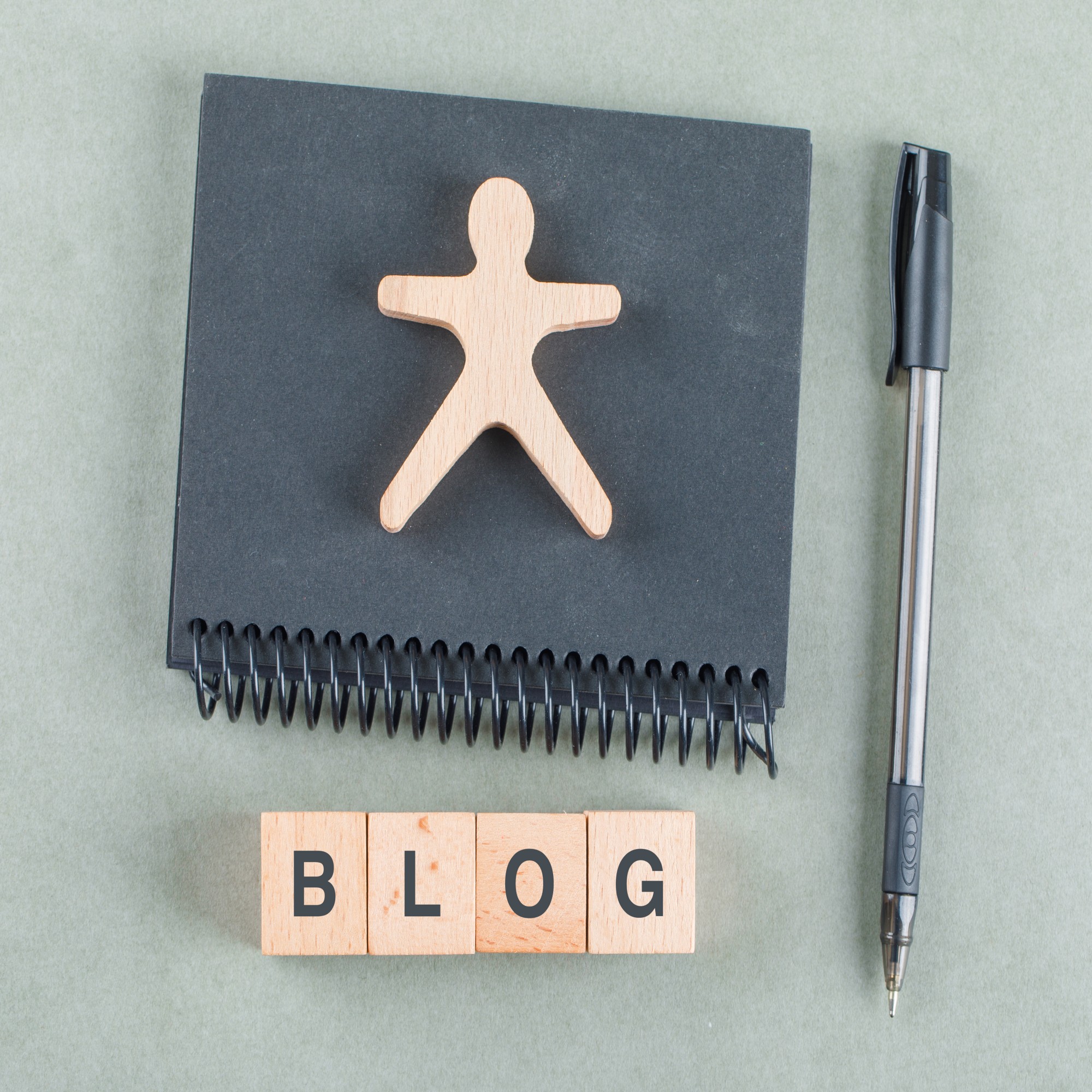 Top view of a notebook, pen, wooden figure, and letter blocks spelling “BLOG” on a desk.